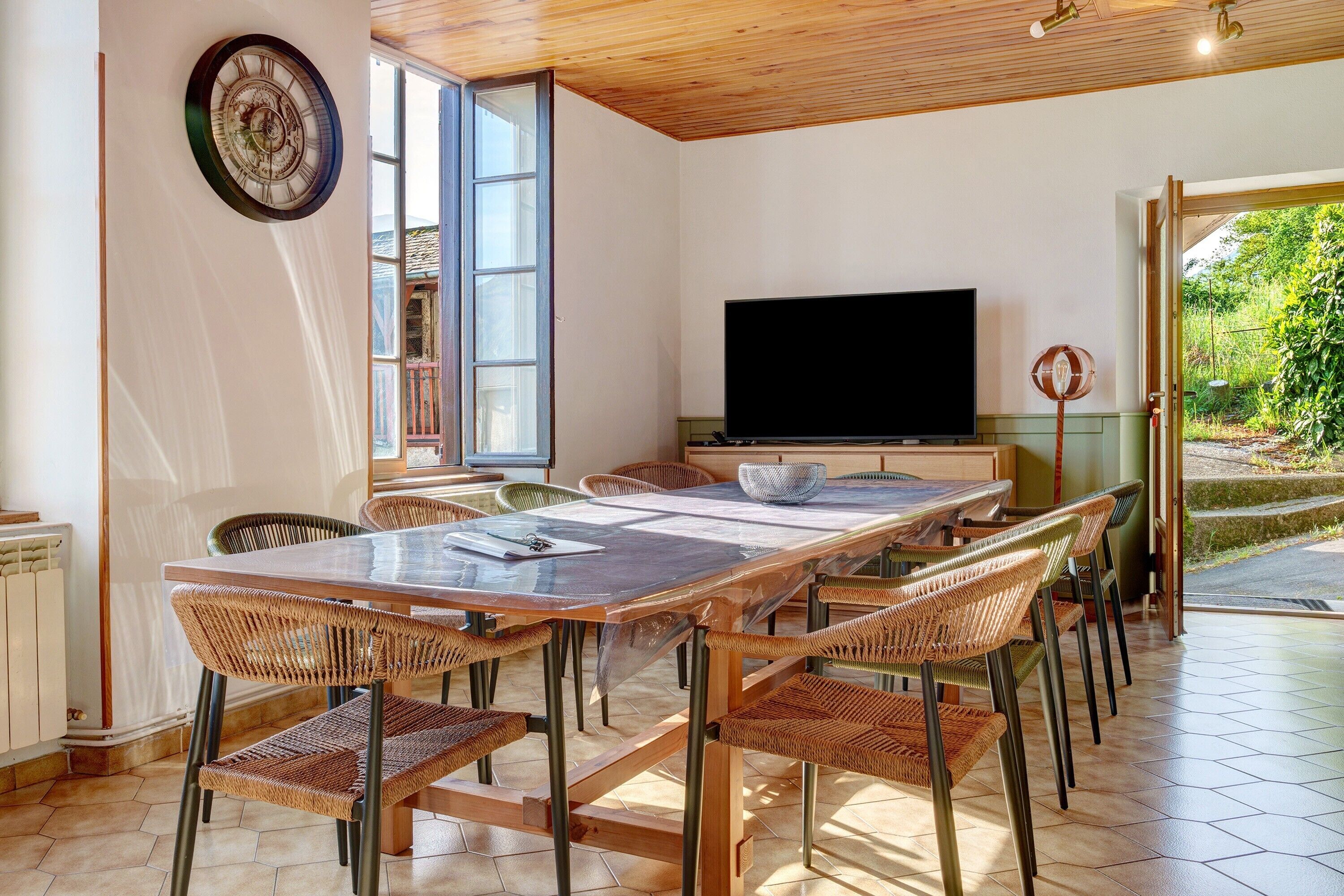 A dining area with a large table, wicker chairs, a flat-screen TV, and a view of greenery outside.