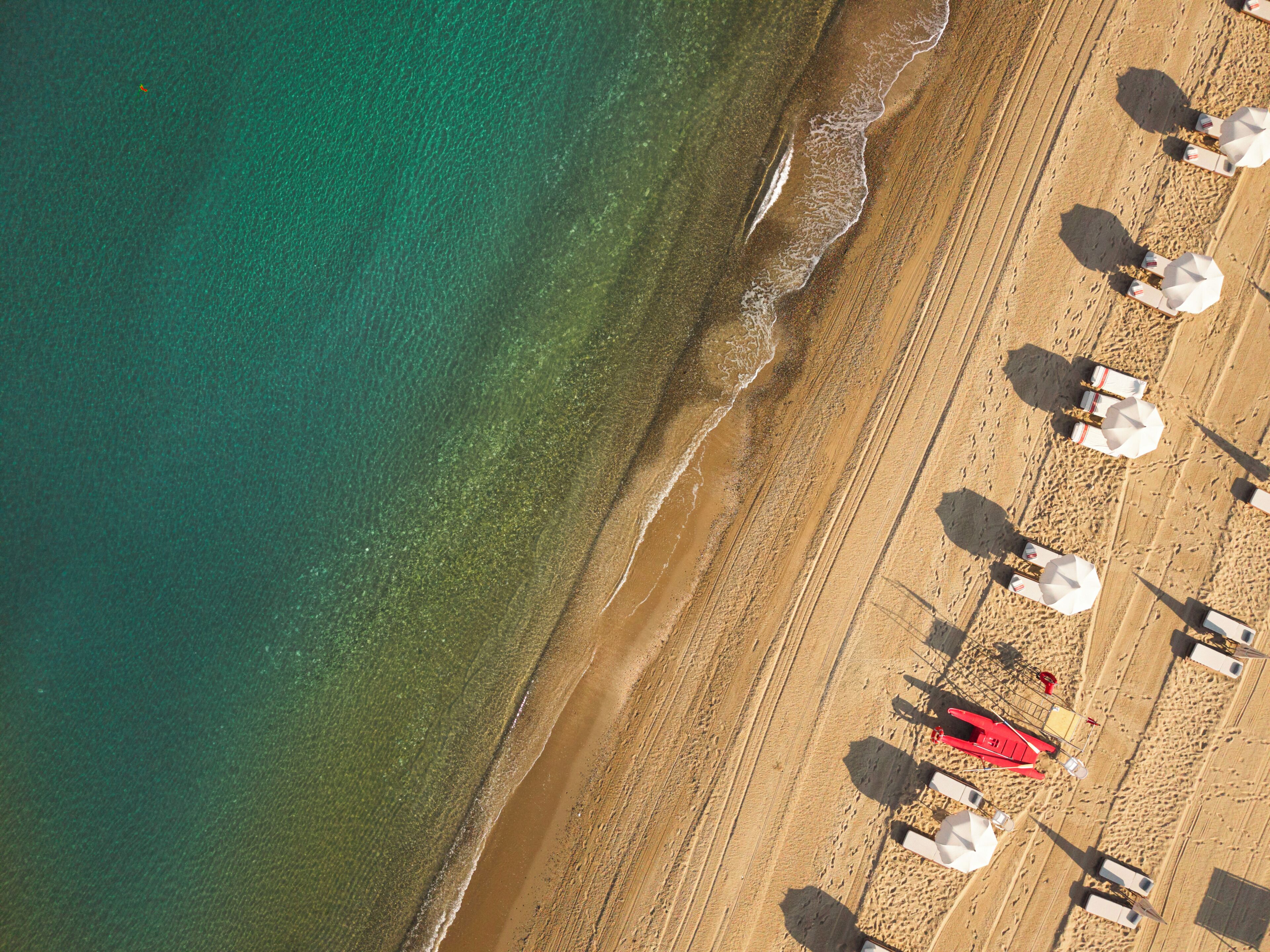 Vista aerea di una spiaggia con file di lettini e ombrelloni, una torretta rossa per i bagnini e un confine netto tra la sabbia e l'acqua.