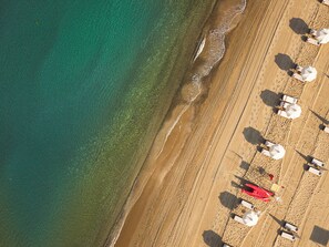An aerial view of a beach with rows of sun loungers and umbrellas, a red lifeguard tower, and a clear demarcation between the sand and the water. -  Icona's Costa Ragusa Resort (Scoglitti)
