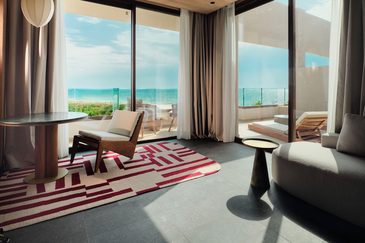 A modern living room with a view of the ocean, a round table, a sofa, and a patterned rug.
