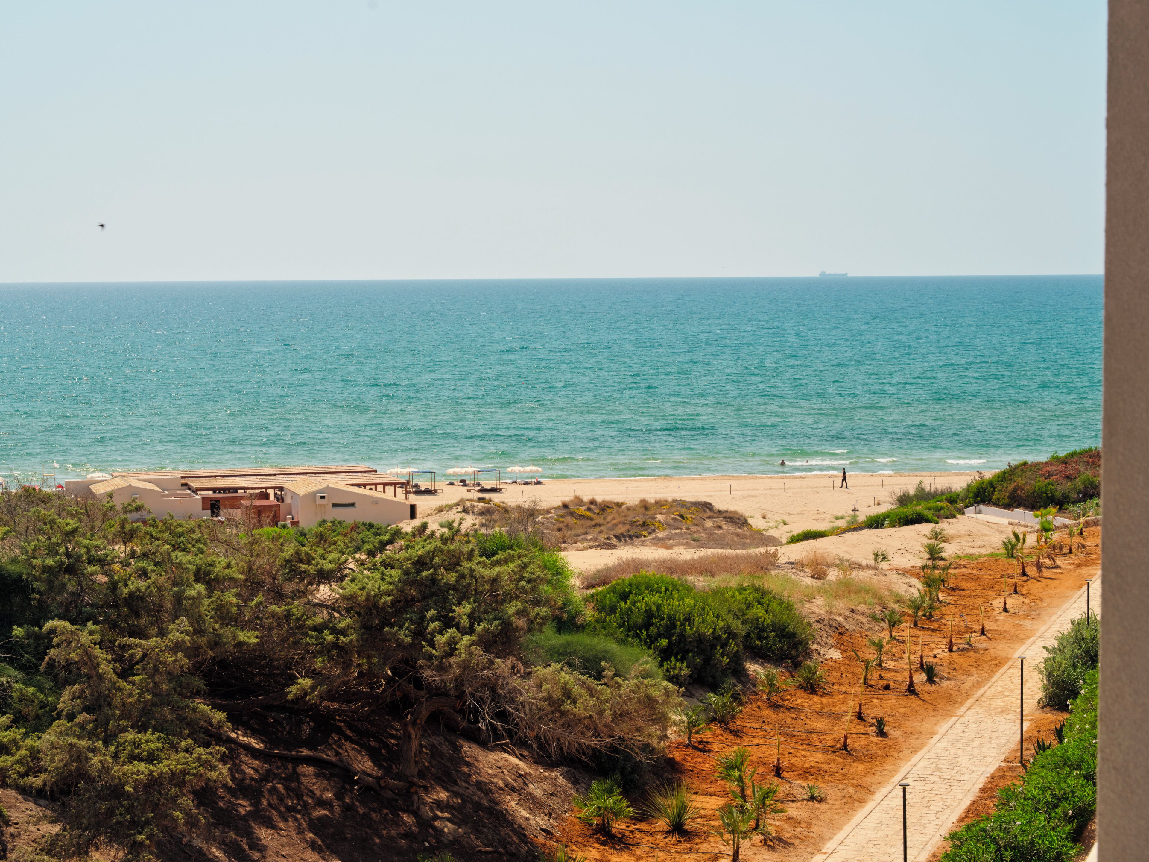 Un paysage côtier avec une plage de sable, une mer bleue et limpide, et un sentier qui traverse un espace aménagé avec des arbustes et de petits arbres.