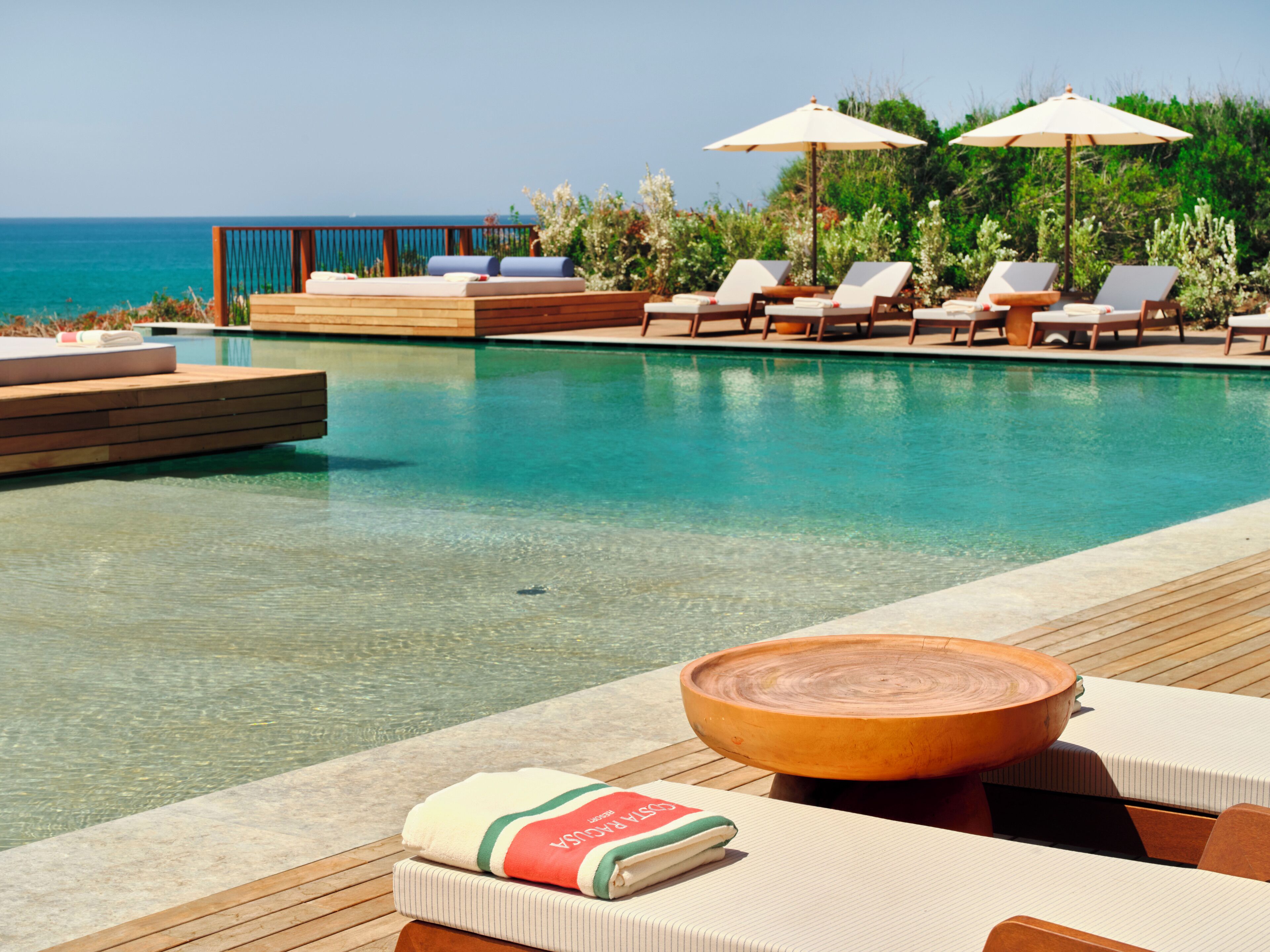 Un espace aménagé au bord de la piscine, avec des chaises longues et des parasols, une terrasse en bois et une vue sur l’océan.