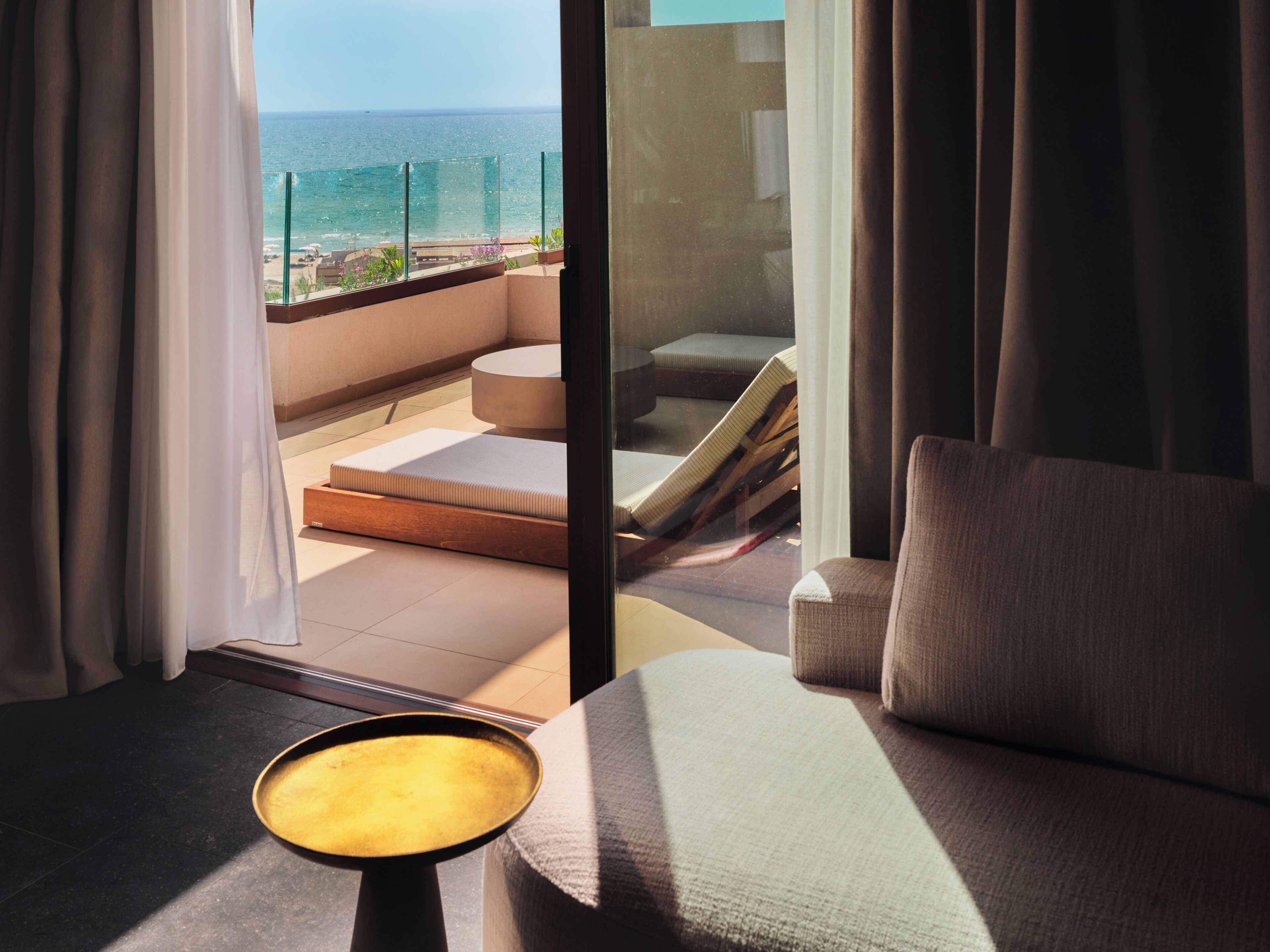 Una habitación con vista al océano, un sofá, una mesita y un ventanal de vidrio que da a un espacio exterior con una banca y una barandilla.