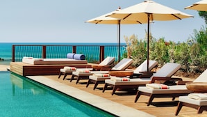 Un espace aménagé au bord de la piscine, avec des chaises longues et des parasols, une terrasse en bois et une vue sur l’océan.