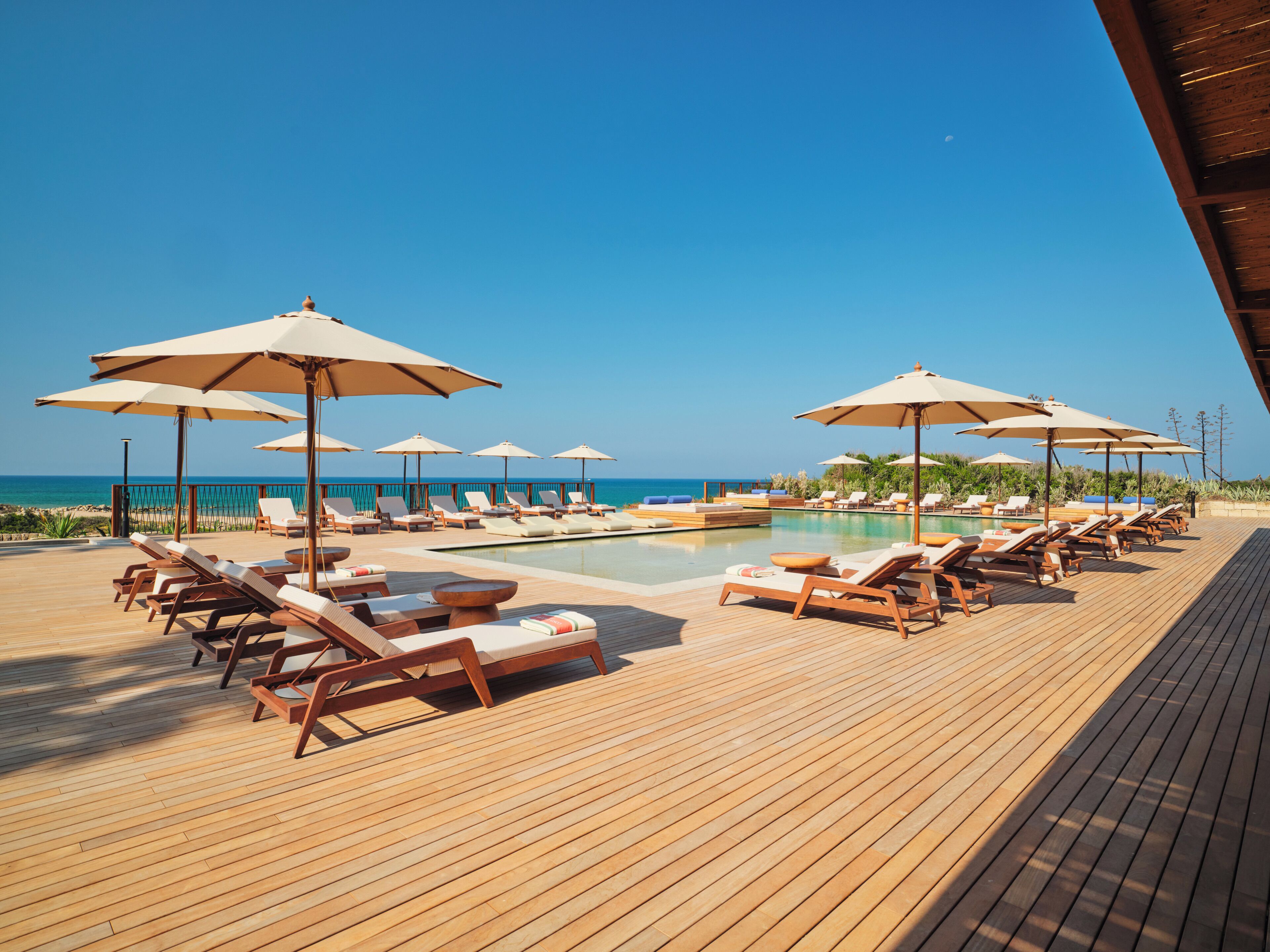 A wooden deck with sun loungers and umbrellas, a pool, and a view of the ocean.