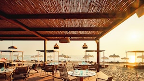 Un espace extérieur aménagé pour les repas, avec des tables et des chaises, des parasols et une vue sur la plage et l’océan.