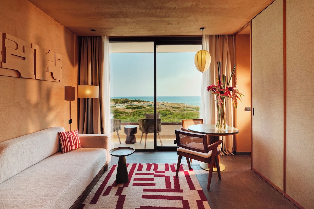 A hotel room with a sofa, a small table, a chair, a view of the beach, and a large window.