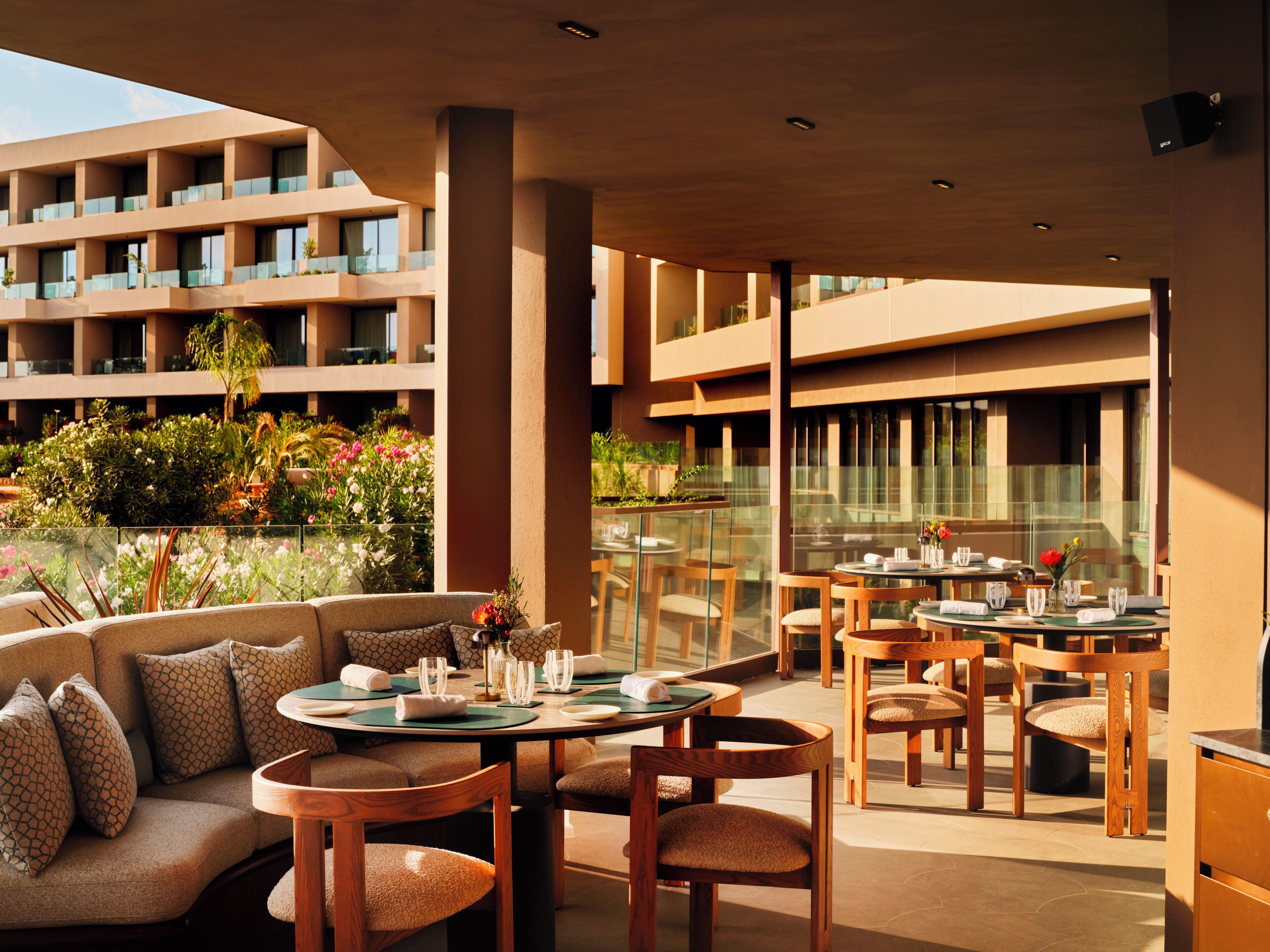 A modern outdoor dining area with round tables and chairs, a sofa, and a view of a building with balconies and greenery.