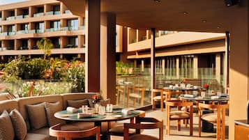 A modern outdoor dining area with round tables and chairs, a sofa, and a view of a building with balconies and greenery.