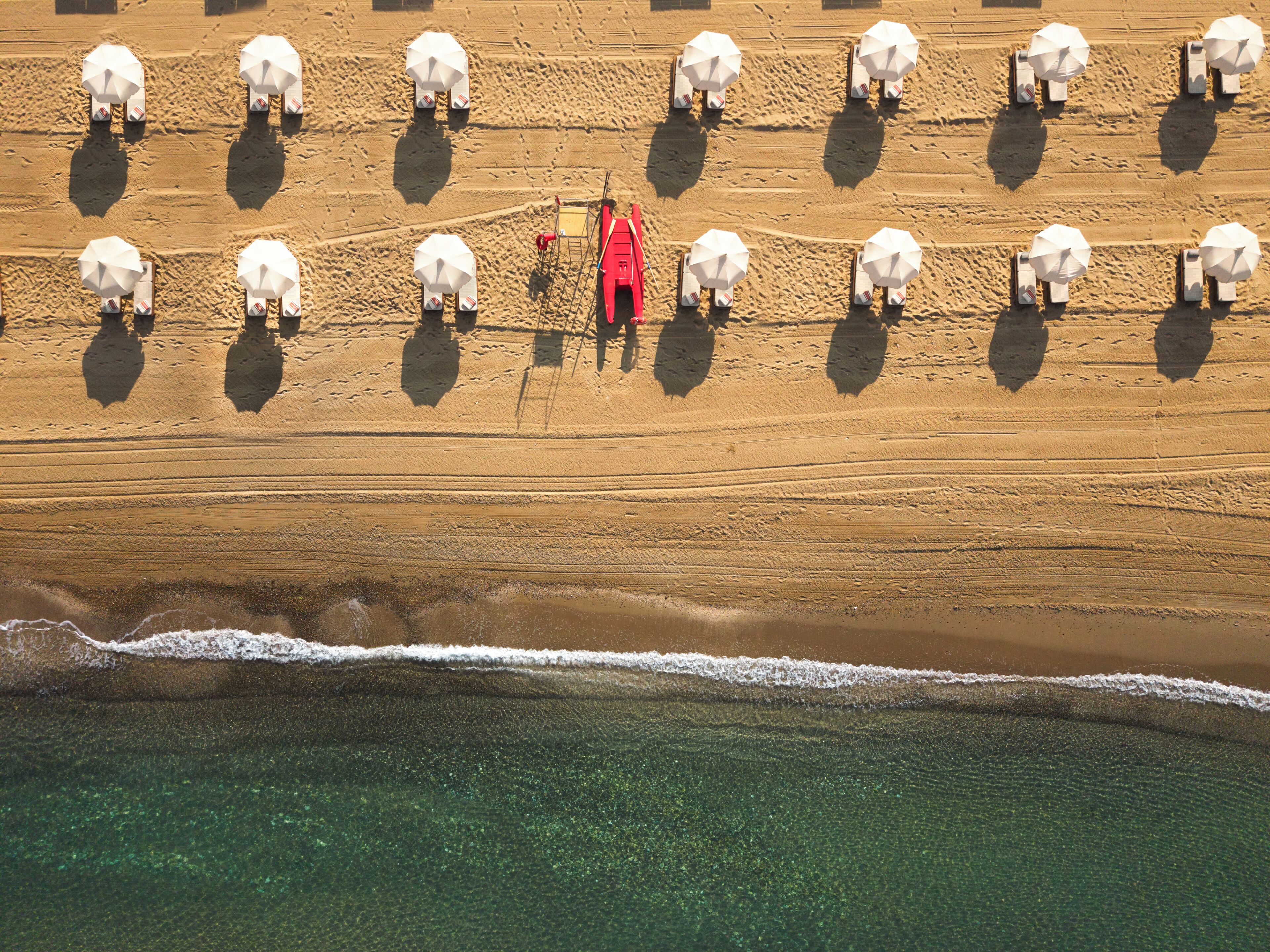 Vue aérienne d’une plage où les parasols sont disposés en rangées, avec un poste de surveillance rouge et l’océan au premier plan.