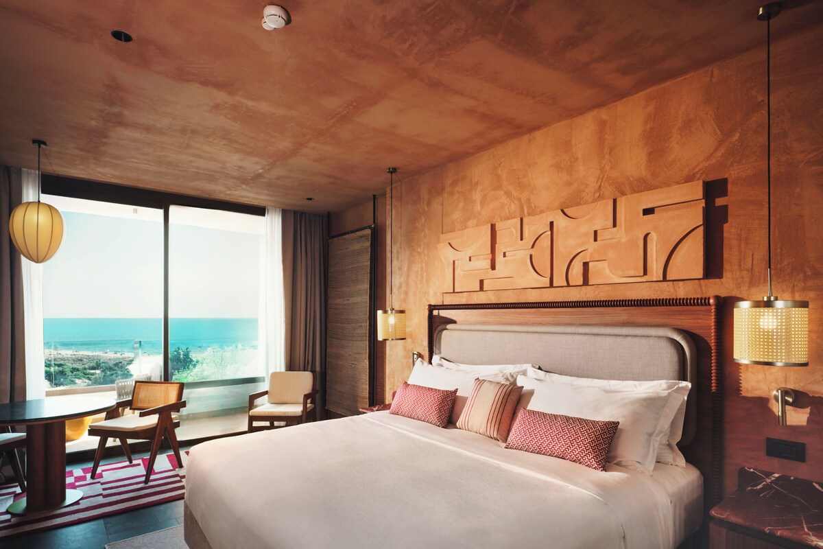 A hotel room with a large bed, a dining area, and a view of the ocean.