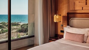 A hotel room with a large window offering a view of the beach and ocean, a bed with pillows, a nightstand, and a wall-mounted lamp. -  Icona's Costa Ragusa Resort - New Opening (Scoglitti)
