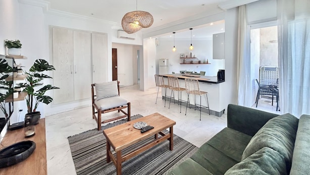 Una sala moderna con un sofá verde, una mesa de centro de madera y un comedor con sillas de mimbre. Se observa una cocina al fondo con armarios blancos y una lámpara colgante.