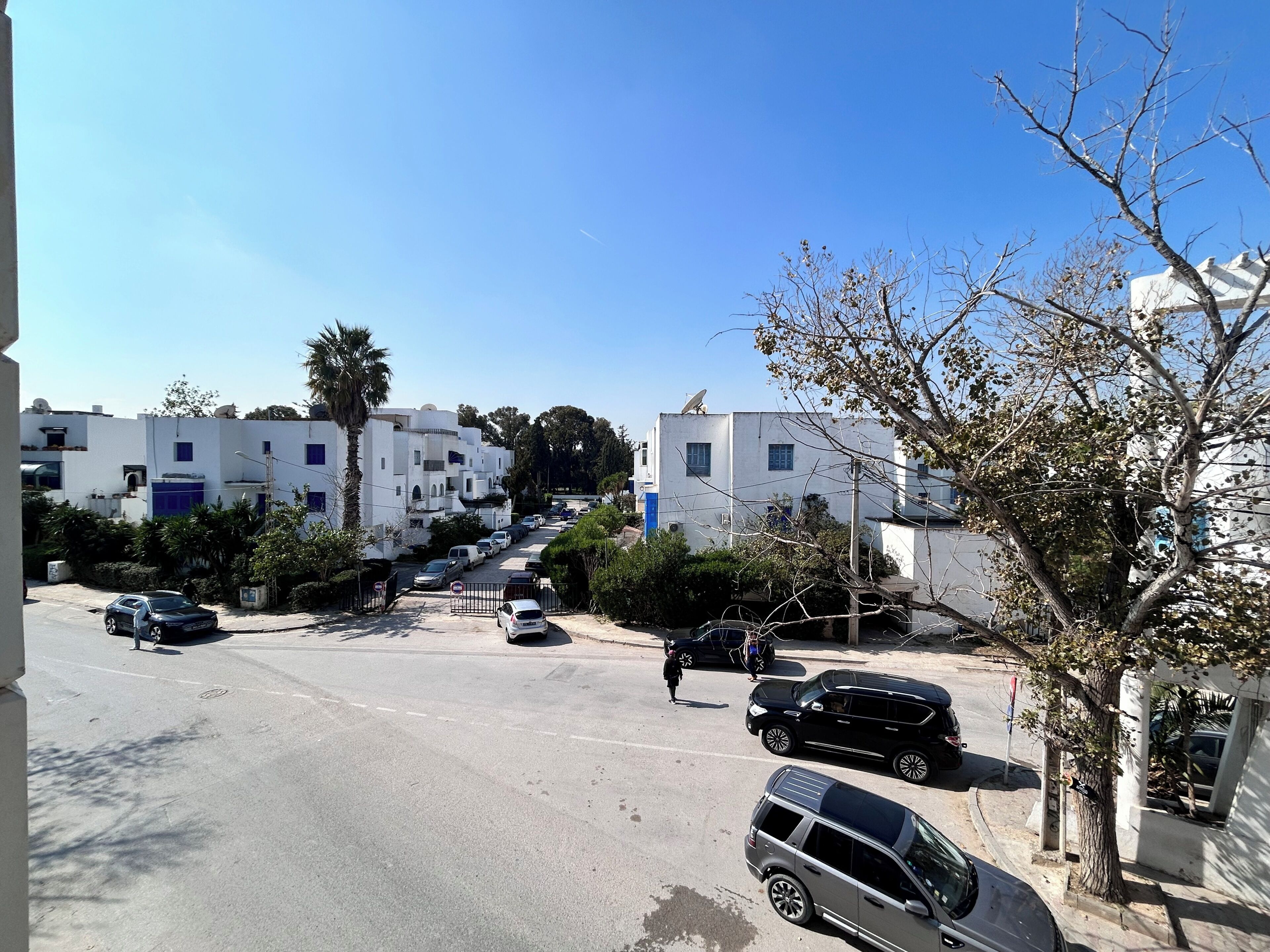 Rua residencial com carros estacionados, edifícios brancos e céu azul claro.