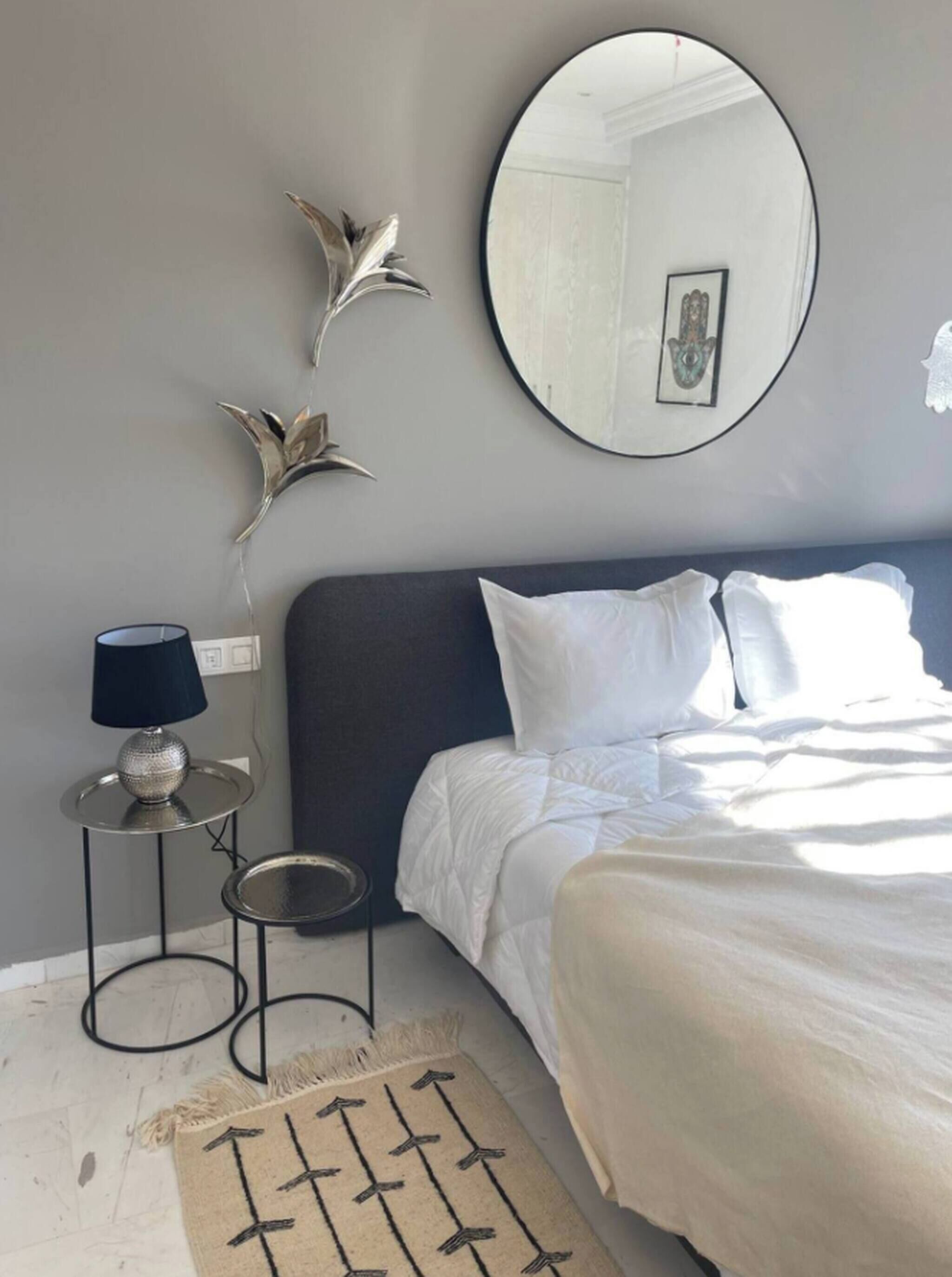 A bedroom with a bed, a round mirror, a small table with a lamp, and decorative wall art.