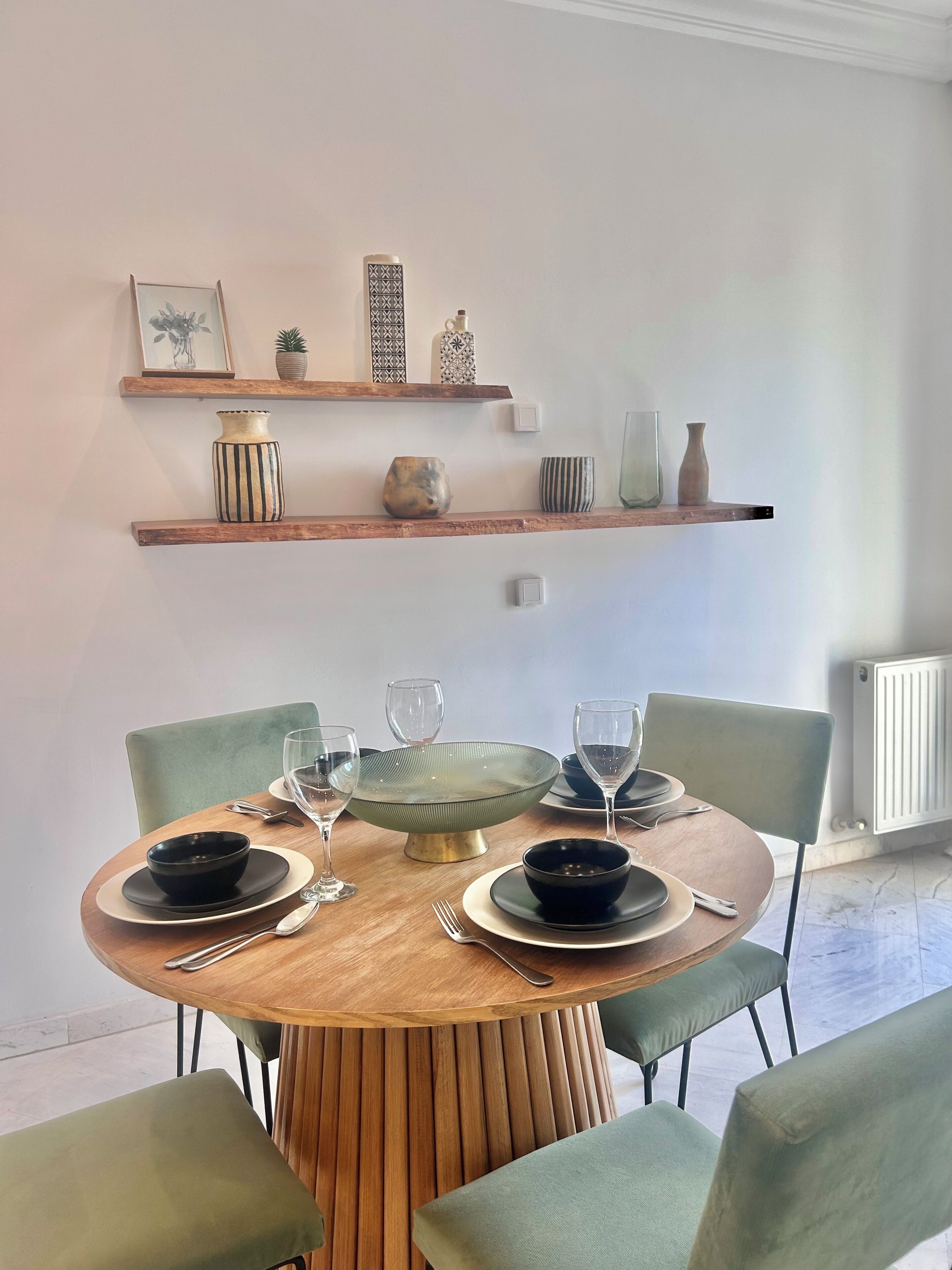 Un comedor con una mesa redonda de madera dispuesta para cuatro personas, rodeada de sillas tapizadas de verde. Encima de la mesa, hay dos estantes de madera con objetos decorativos.