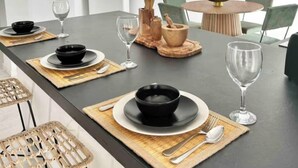 Un comedor moderno con una larga mesa dispuesta para dos personas, con cuencos, platos y cubiertos negros, además de un cuenco de madera y una piña sobre la mesa.