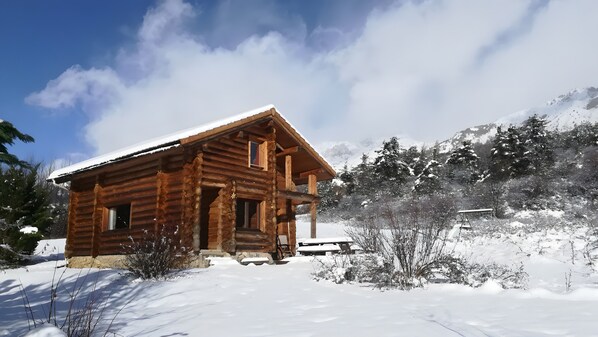 Une cabane en bois dont le toit est recouvert de neige, entourée d’arbres et de montagnes également recouverts de neige.