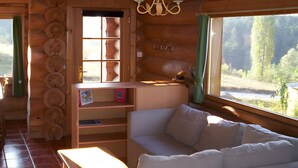 A log cabin interior with a sofa, a table, a bookshelf, and a window with a view of a forest. - Le chalet neige – Chorges Hautes-Alpes (Chorges)