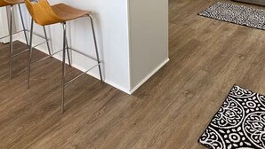 Eine moderne Küche mit weißen Schränken, einer schwarzen Arbeitsplatte und Edelstahlgeräten. Zwei hölzerne Barhocker und ein gemustertes Teppich sind auf dem Boden zu sehen.