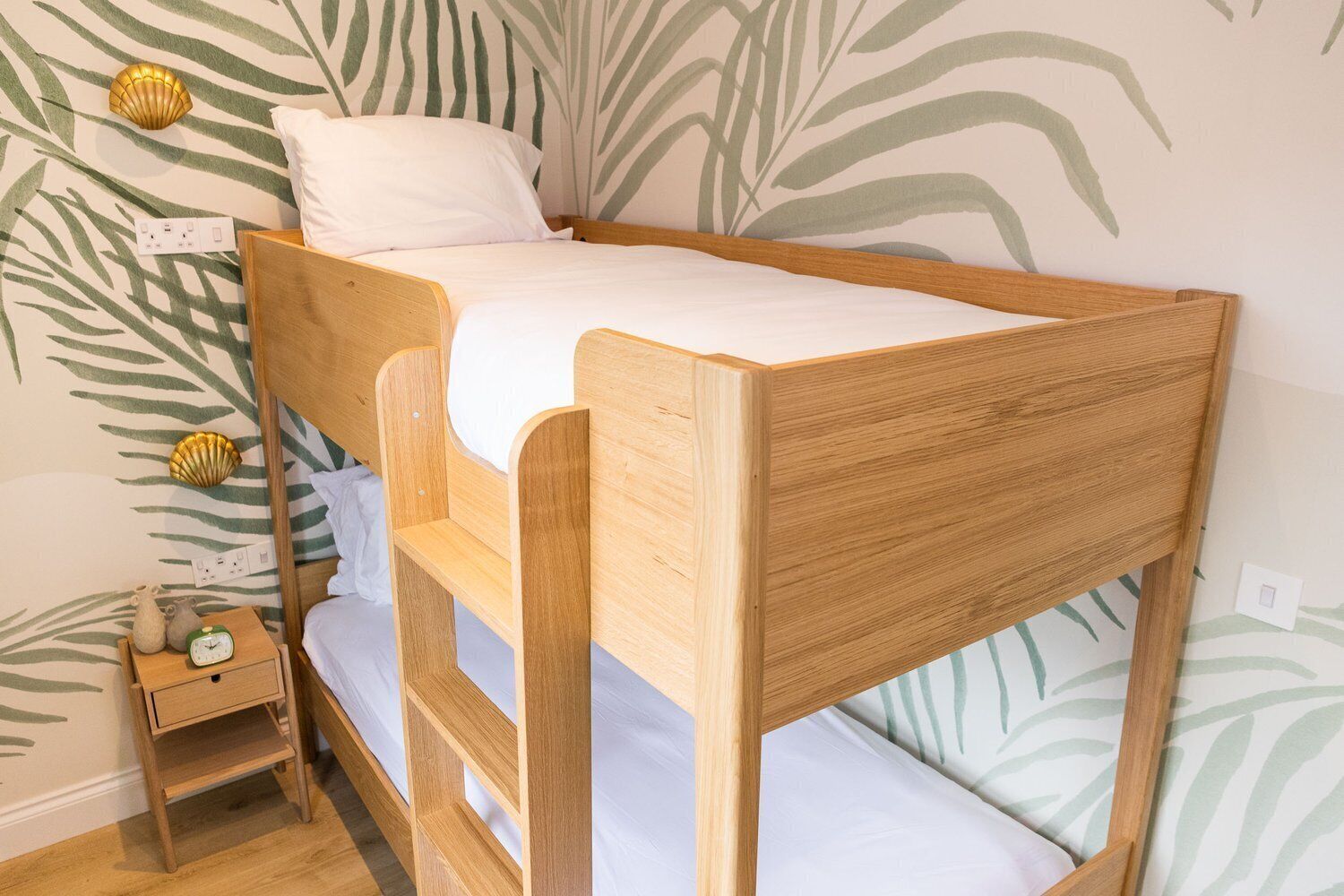 A bunk bed with a wooden frame and white bedding, a small bedside table with a clock, and a wall with a palm leaf pattern.