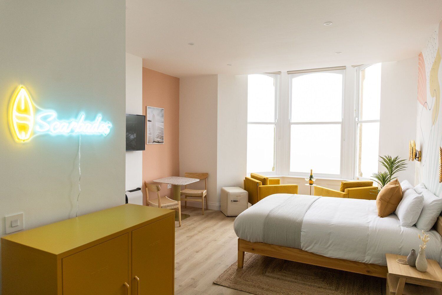A modern hotel room with a bed, a desk, a chair, a small table, and a neon sign.