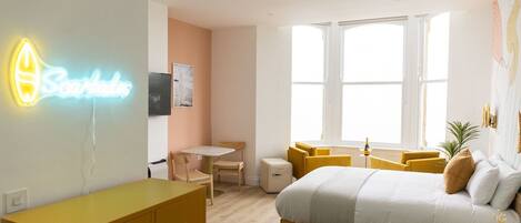 A modern hotel room with a bed, a desk, a chair, a small table, and a neon sign.