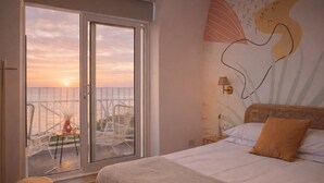 A bedroom with a bed, a nightstand, a wall lamp, a window with a view of the sunset, and a balcony with chairs and a table. - Beach House Rooms (Scarborough)