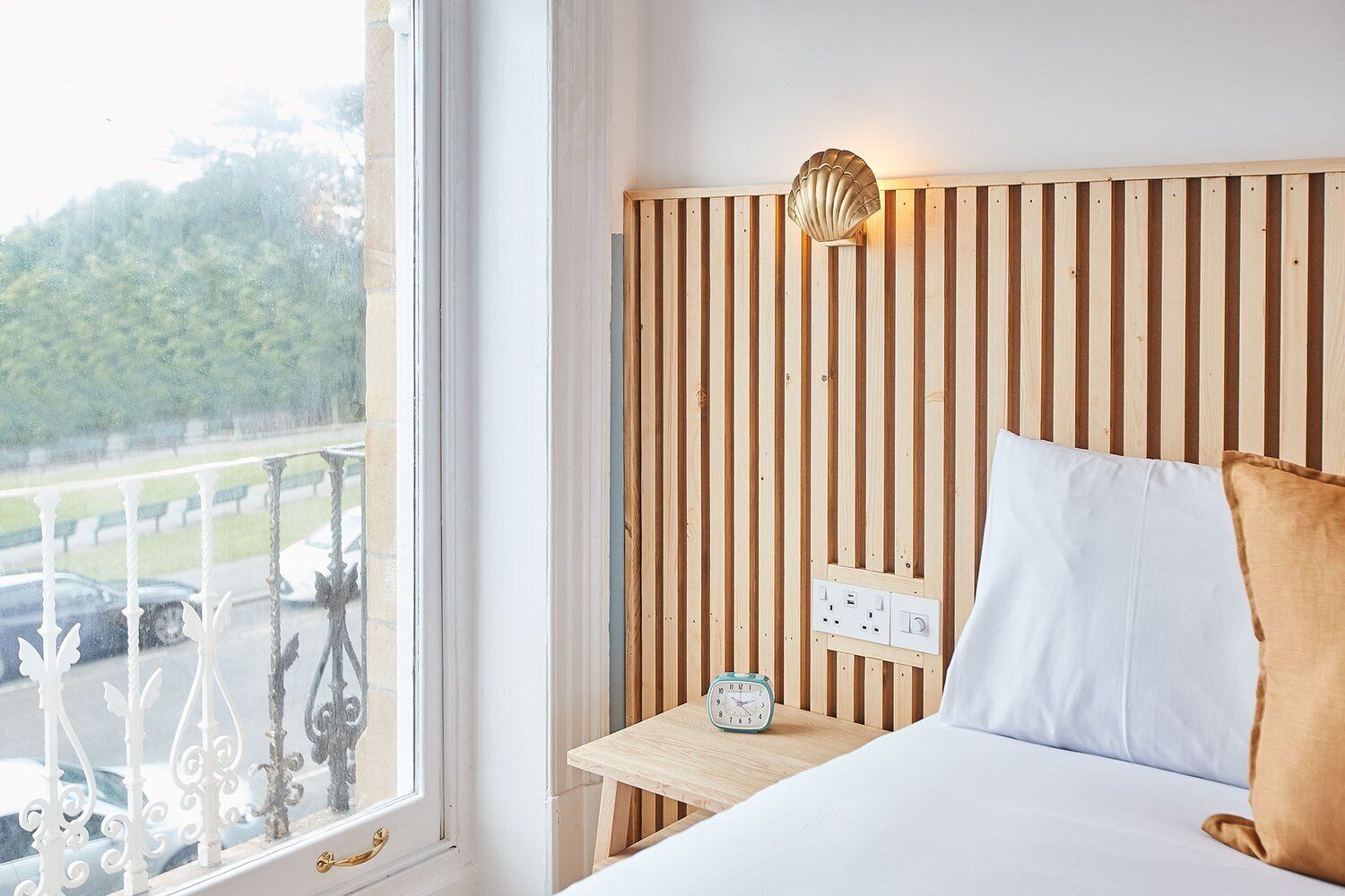 A hotel room with a wooden slat headboard, a bedside table with a clock, and a view of a parking area through a window with ornate white railings.