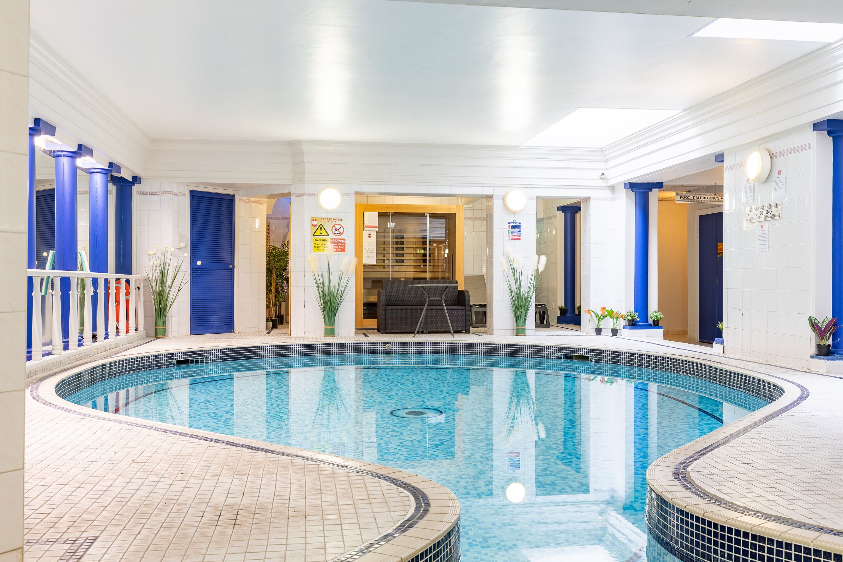 A swimming pool with a tiled deck, blue pillars, and a lounge area with a sofa and table.