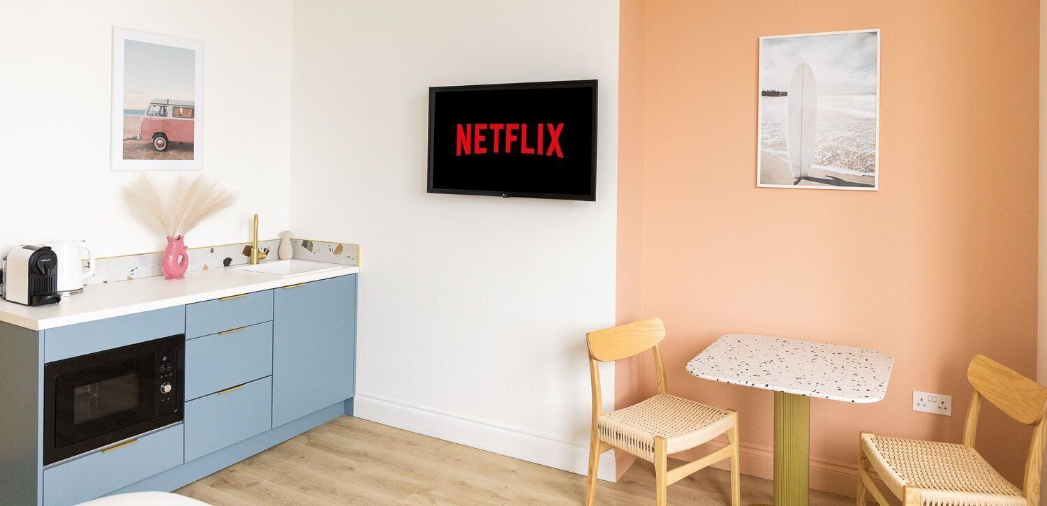 A modern kitchen with blue cabinets, a microwave, a coffee maker, and a pink vase. A TV screen displays "NETFLIX" in red letters.