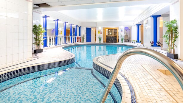 An indoor swimming pool with tiled walls and blue pillars, surrounded by potted plants and a tiled floor. - Beach House Rooms (Scarborough)