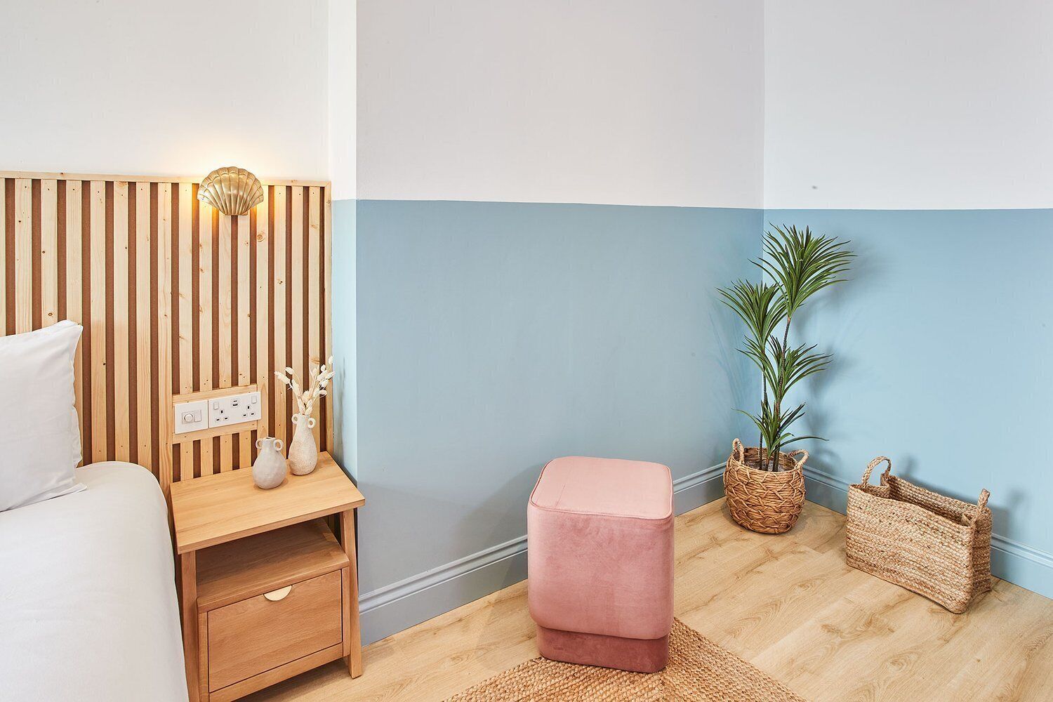 A bedroom with a wooden slat headboard, a bedside table with a lamp, a pink ottoman, a potted plant, and a woven basket.