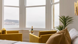 A room with a bed, two yellow armchairs, a small table with a bottle and glasses, and a large window with a view of a distant city. - Beach House Rooms (Scarborough)