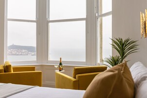 Una habitación con una cama, dos sillones amarillos, una mesita con una botella y vasos, y un ventanal con vistas a una ciudad distante.