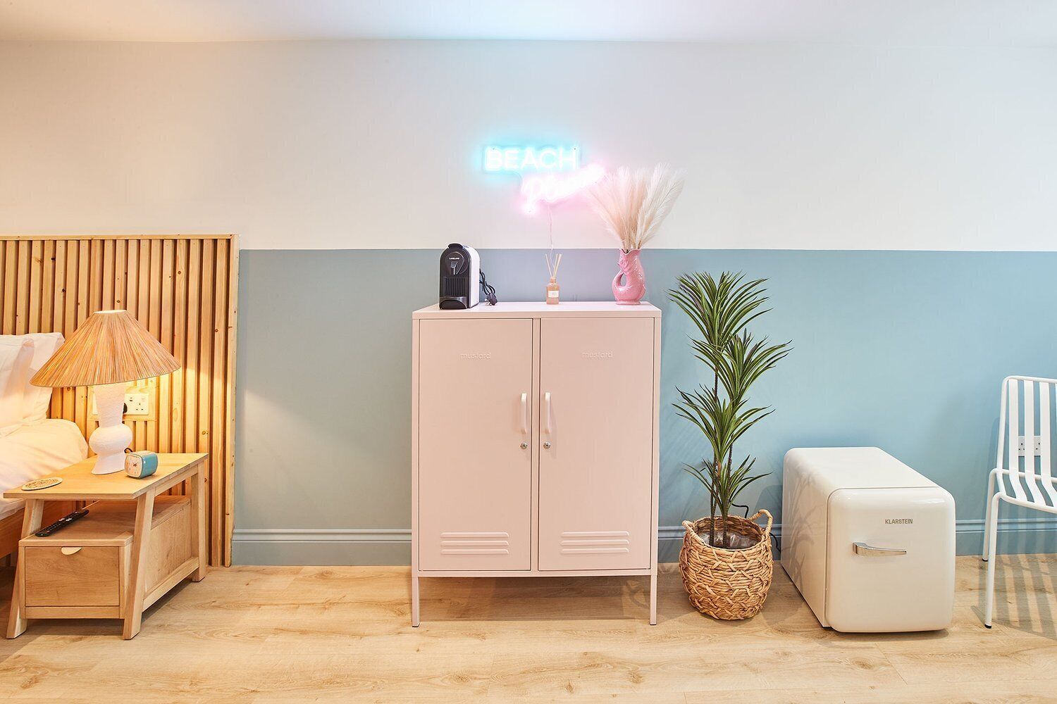 A room with a wooden slat wall, a pink cabinet, a small table with a lamp, a plant, and a vintage refrigerator.