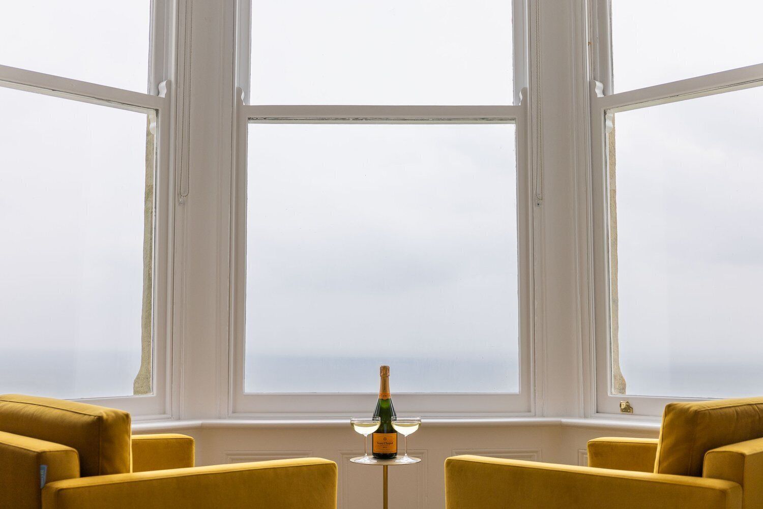 A room with a large window, two yellow chairs, and a table with a bottle and glass.