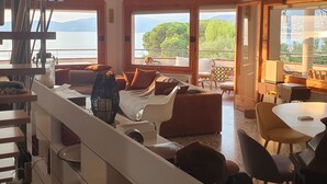 Amplia sala de estar con un ventanal que ofrece vistas al mar y a las colinas. Hay un sofá, una mesa de comedor y un ventilador de techo.