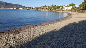 Una playa de guijarros con un mar tranquilo, edificios blancos y árboles verdes al fondo.