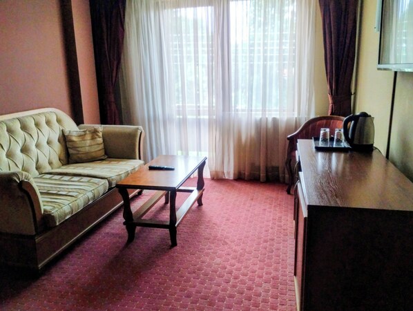 A furnished room with a sofa, a coffee table, a desk with a kettle, and a chair. - Hotel Sofia (Bansko)