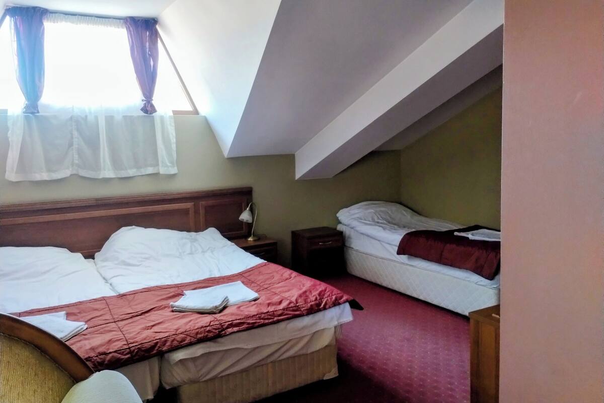 A hotel room with two beds, a wooden headboard, a nightstand, a lamp, and a window with curtains.