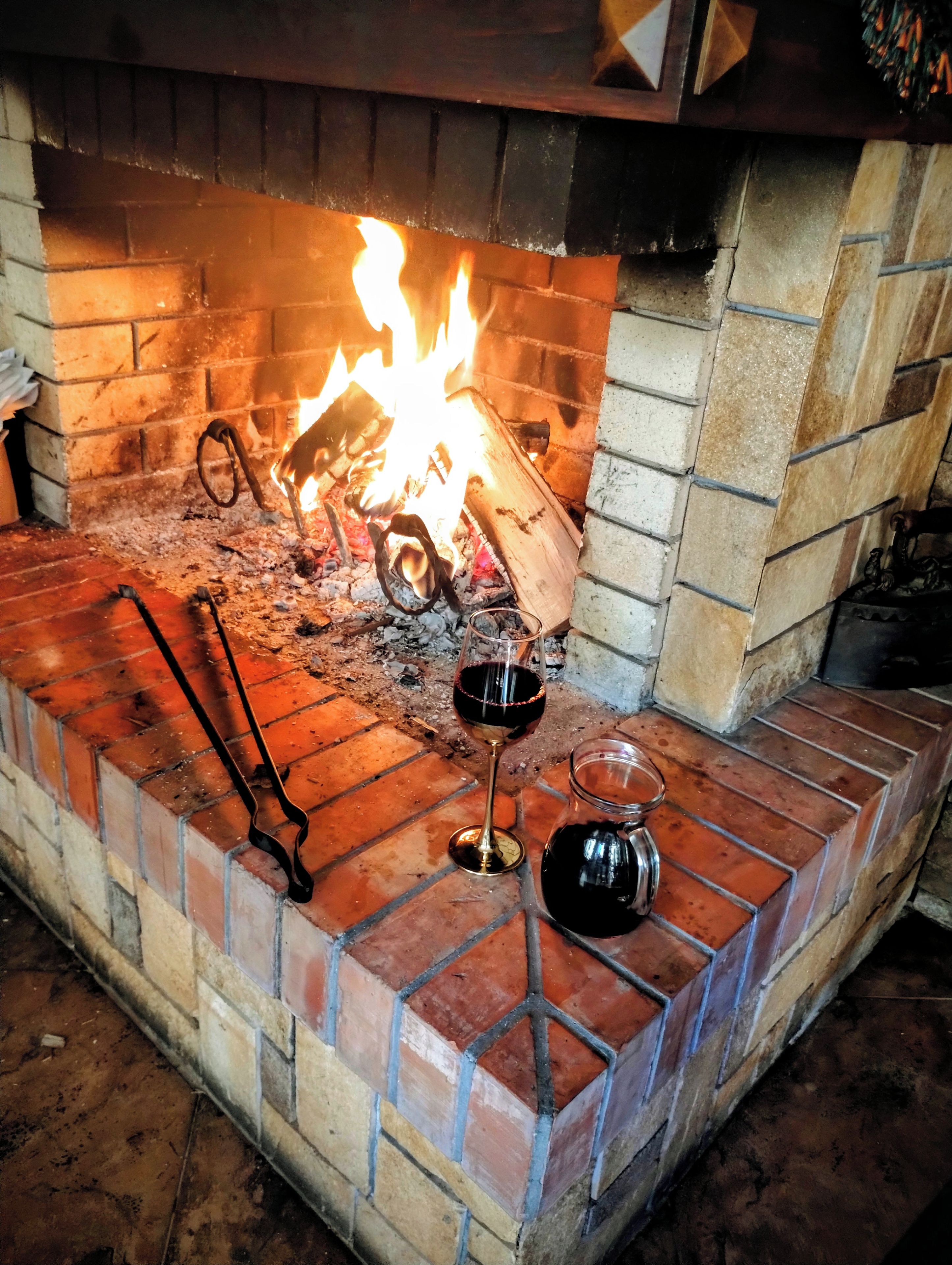 A fireplace with a fire, a glass of red wine, and a metal pitcher on a brick hearth.