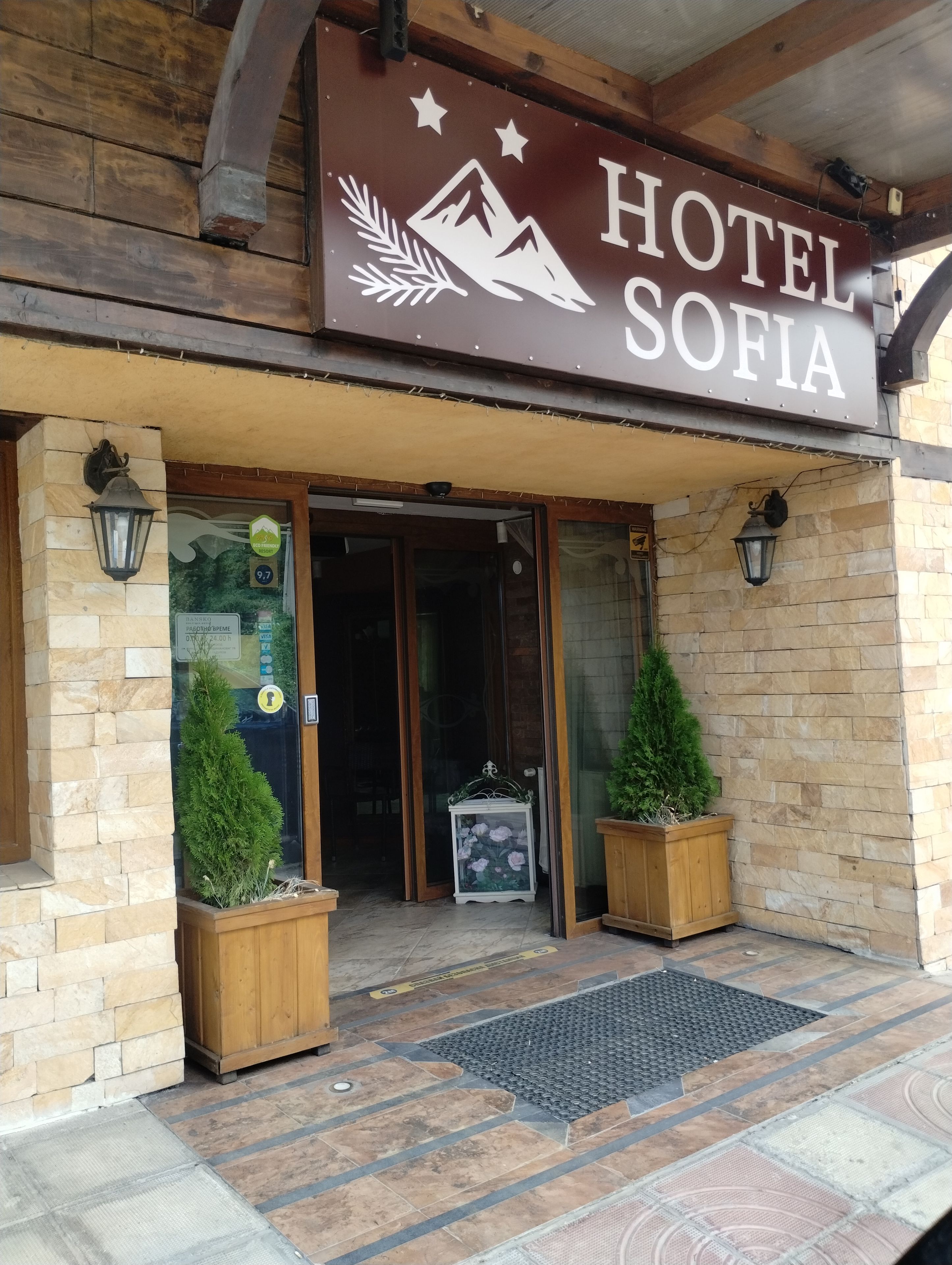 A hotel entrance with a sign reading 'HOTEL SOFIA,' a wooden facade, and two potted plants at the entrance.