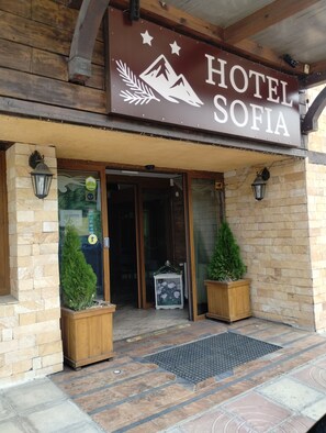 A hotel entrance with a sign reading 'HOTEL SOFIA,' a wooden facade, and two potted plants at the entrance. - Hotel Sofia (Bansko)