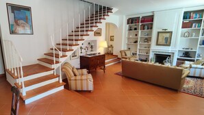 Amplia sala de estar con una escalera, chimenea y estantería llena de libros.
