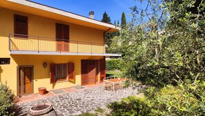 Un edificio amarillo de dos pisos con contraventanas de madera, un patio empedrado en piedra y abundante vegetación.