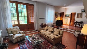 Un salón con sofá, sillón, mesa de centro y una lámpara. Hay una chimenea, una estantería y una ventana con cortinas.