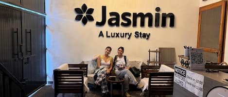 A reception area with a sign for 'Jasmin A Luxury Stay,' a chandelier, and a notice board with check-in/out times.
