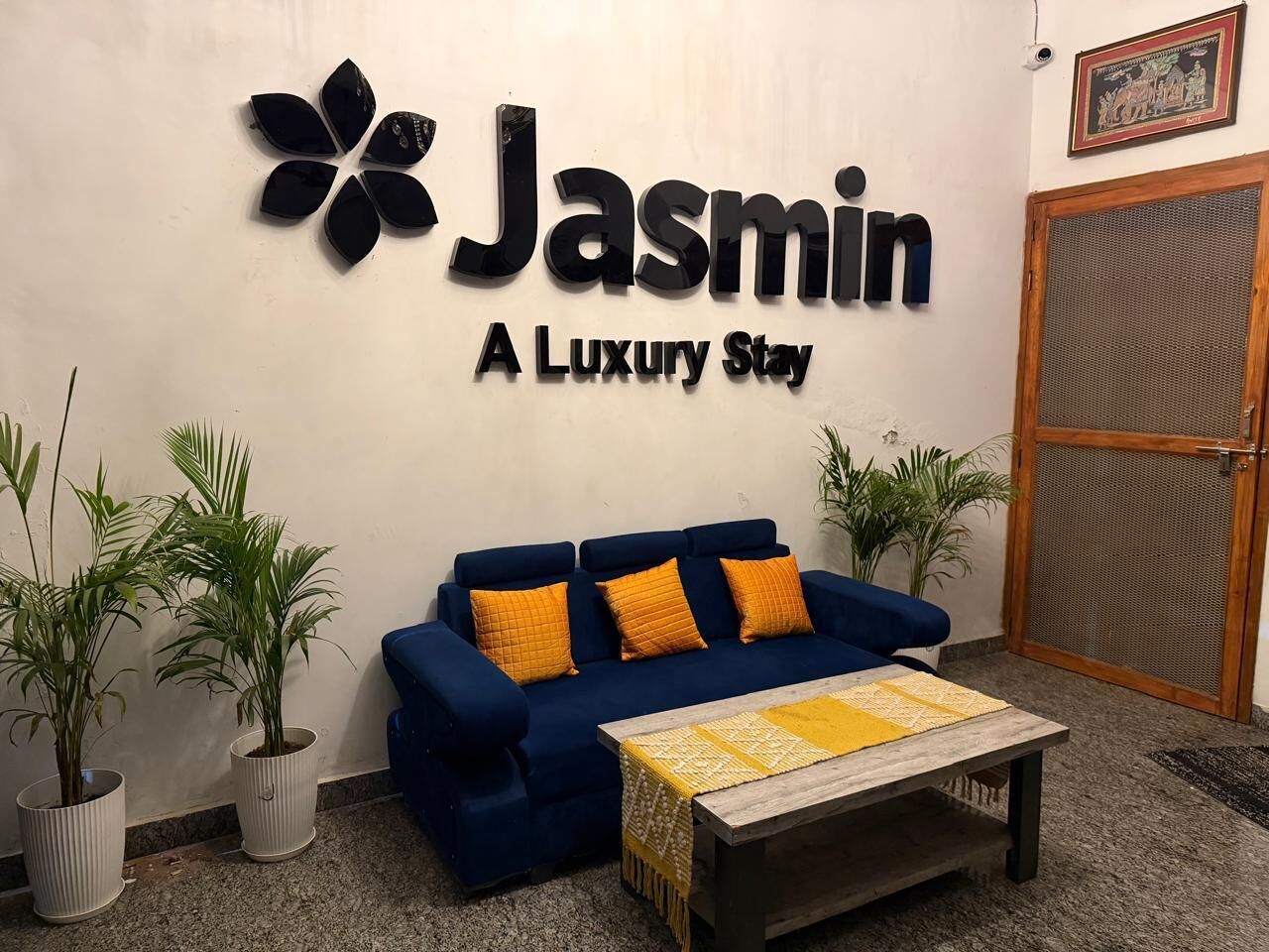 Egy tágas előtér, melyben egy kék kanapé, két virágtálban lévő növény és egy „Jasmin – luxus szállás” feliratú tábla található.