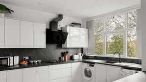 A modern kitchen with white cabinets, a black countertop, a built-in oven, a microwave, a sink, and a window with a view of trees. - Hartley Avenue (Leeds)